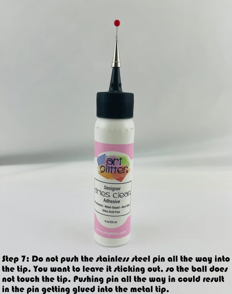 A bottle of Art Glitter Designer dries Clear adhesive shown on a light gray background with a black text overlay at the bottom explaining how to handle the stainless steel pin tip.