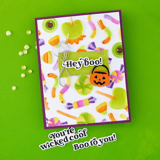 A handmade Halloween greeting card with a white background covered in colorful candy illustrations, a green 'Hey boo!' label, and a small jack-o'-lantern. The card is displayed on a bright green background with decorative rhinestones and the text 'You're wicked cool Boo to you!' below it.
