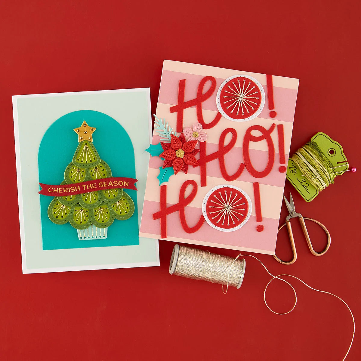 Two handmade greeting cards displayed on a red background with crafting props including scissors, thread, and a spool of green thread.