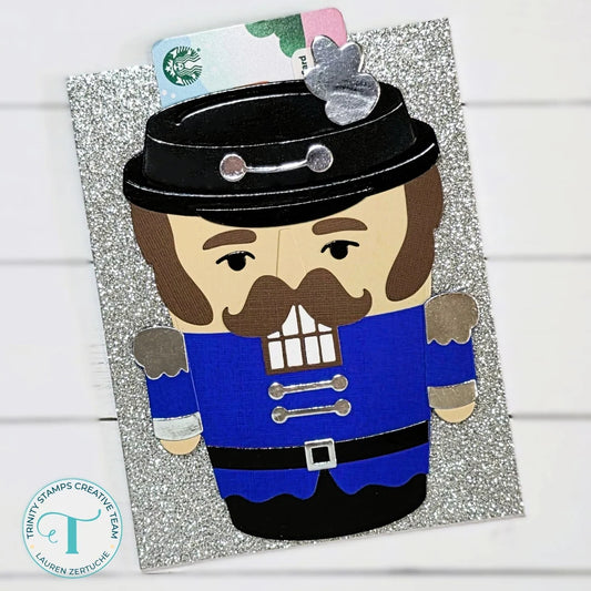 A Christmas nutcracker card featuring a paper-cut nutcracker in a blue coat and black hat on a glittery silver background, with a Starbucks card tucked behind the head and a small silver ornament.