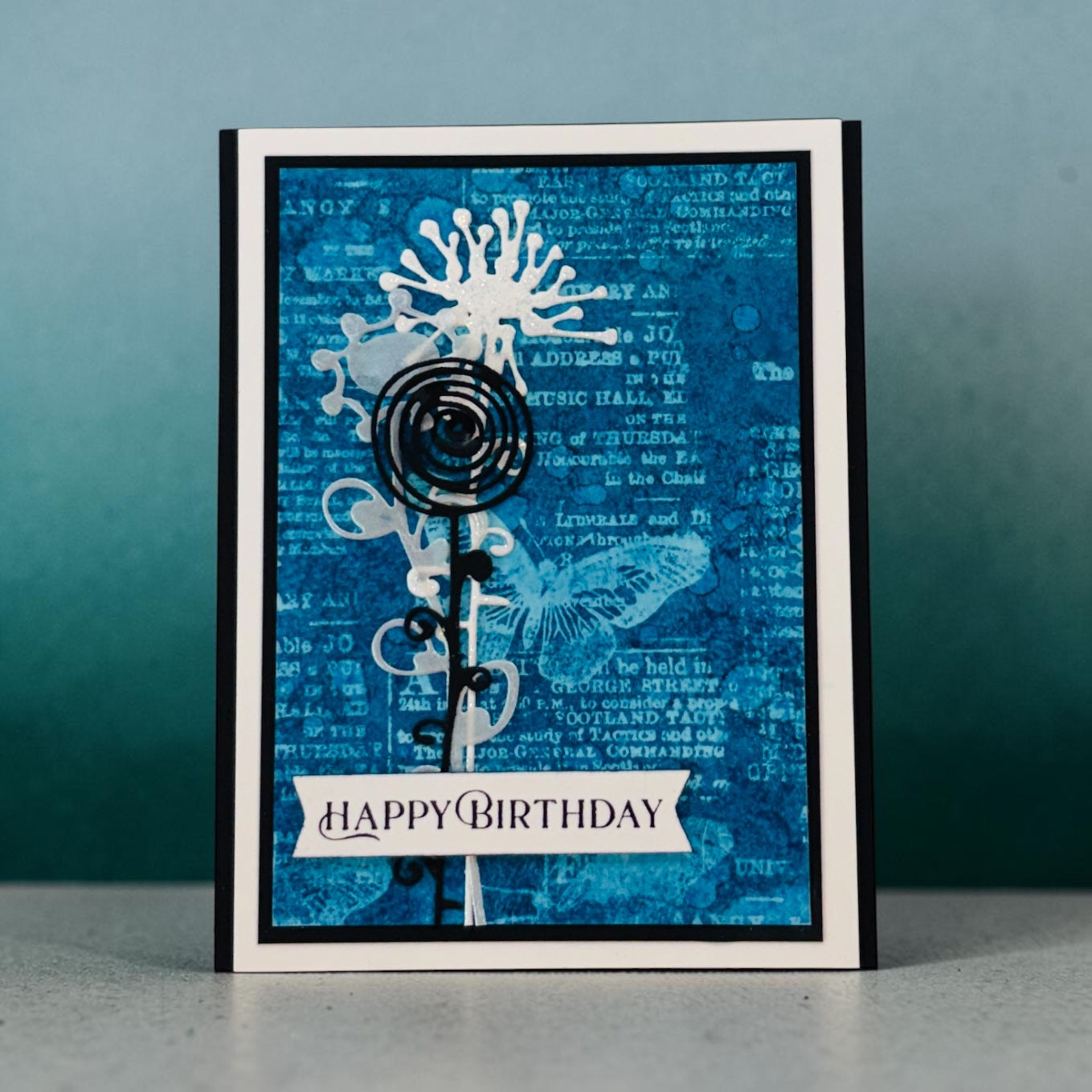 A handmade greeting card with a blue abstract background, layered black and white floral elements, and a white banner reading 'HAPPY BIRTHDAY', photographed standing upright against a blue wall.