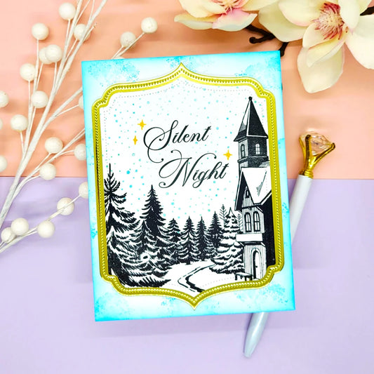 A square handmade card with a winter church scene and the words 'Silent Night' in black script, styled on a pastel pink and lavender surface with flowers and a pen nearby.