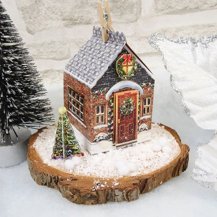 A decorative miniature paper Christmas cottage ornament displayed on a rustic wood slice base with faux snow, a small lit tree, and a white fabric backdrop.