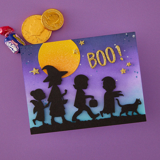 A Halloween-themed square card or art print with black silhouettes of children and a cat walking under a large yellow moon and purple-to-blue gradient sky, with gold glitter stars and the word 'BOO!' in gold. The item is photographed on a purple background with two gold coins and a wrapped candy placed beside it as props.