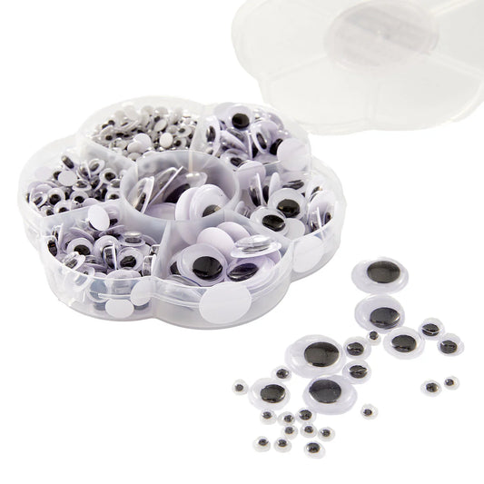 A clear plastic compartment organizer filled with white and black plastic googly eyes, with several loose googly eyes scattered on a white background. A translucent lid is partially visible at the top right.