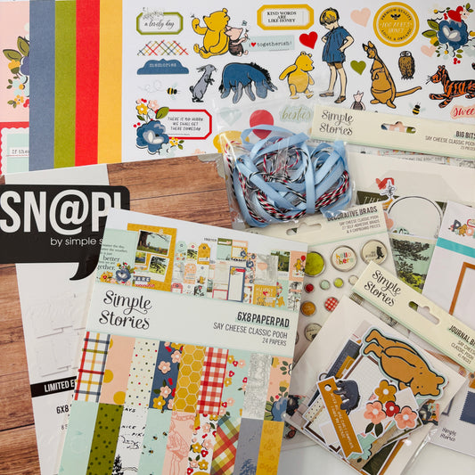 Scrapbooking supplies including paper pads, stickers, and ribbons on a wooden surface with 'Simple Stories' branding.