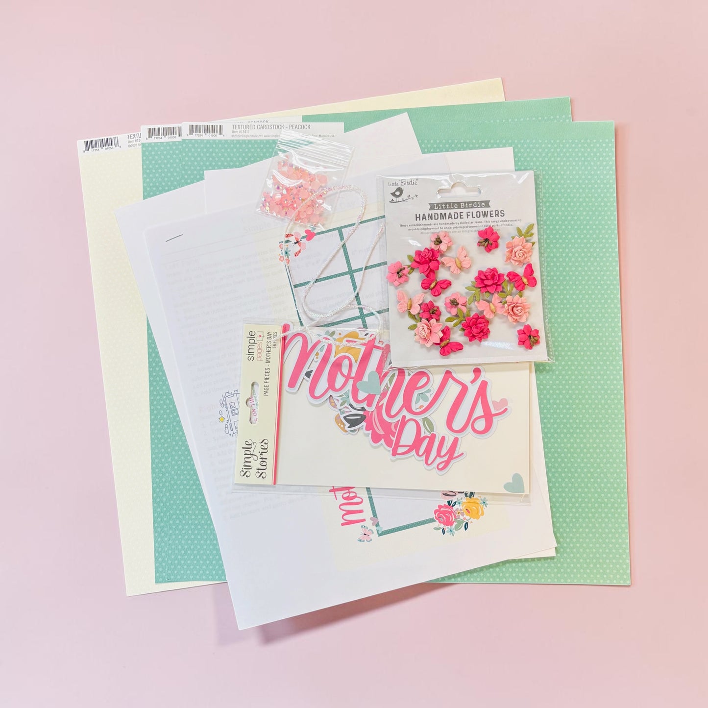 A flat-lay arrangement of Mother's Day themed craft supplies, including a clear stamp with floral designs, a sheet of pink 'Mother's Day' script, various floral embellishments, and patterned paper sheets on a soft pink background.