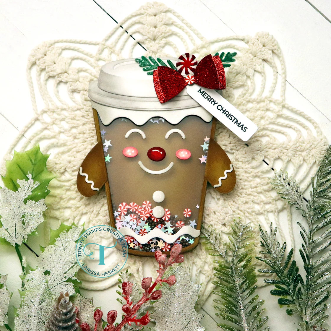 A festive gingerbread coffee cup decoration with a smiling face, red nose, white icing details, a red glittery bow, and a 'MERRY CHRISTMAS' tag, styled on a white crochet blanket with holiday greenery.