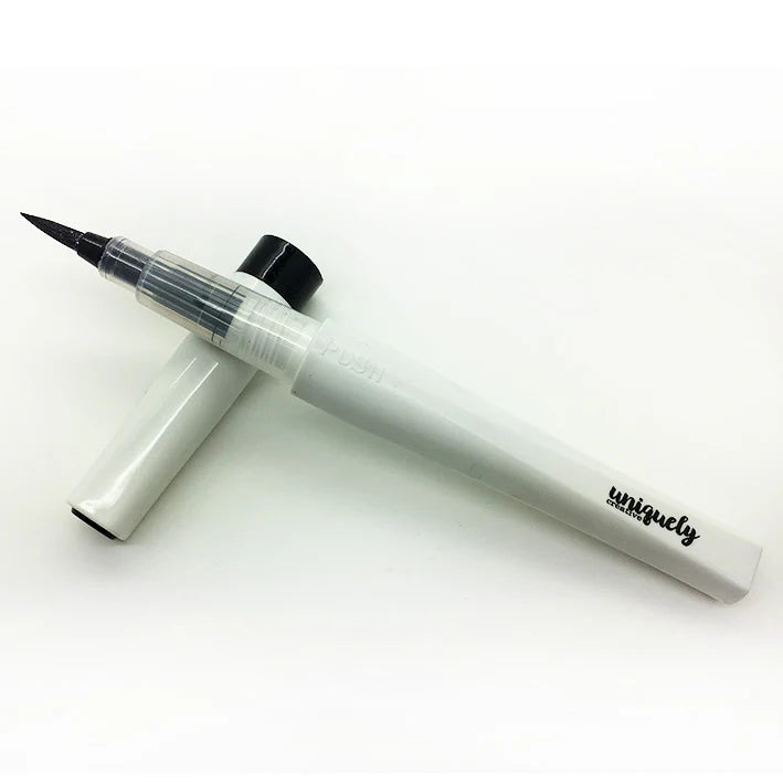 A white and clear brush-tip pen with a black cap, shown with its cap removed and placed behind it on a white background. The handle has the word 'uniquely' printed in black script.