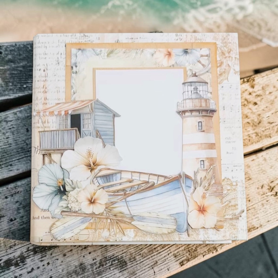 A square decorative scrapbook or photo album cover featuring a nautical beach theme with a lighthouse, flowers, a small boat, and a blank photo frame, displayed on weathered wooden boards with ocean water in the background.