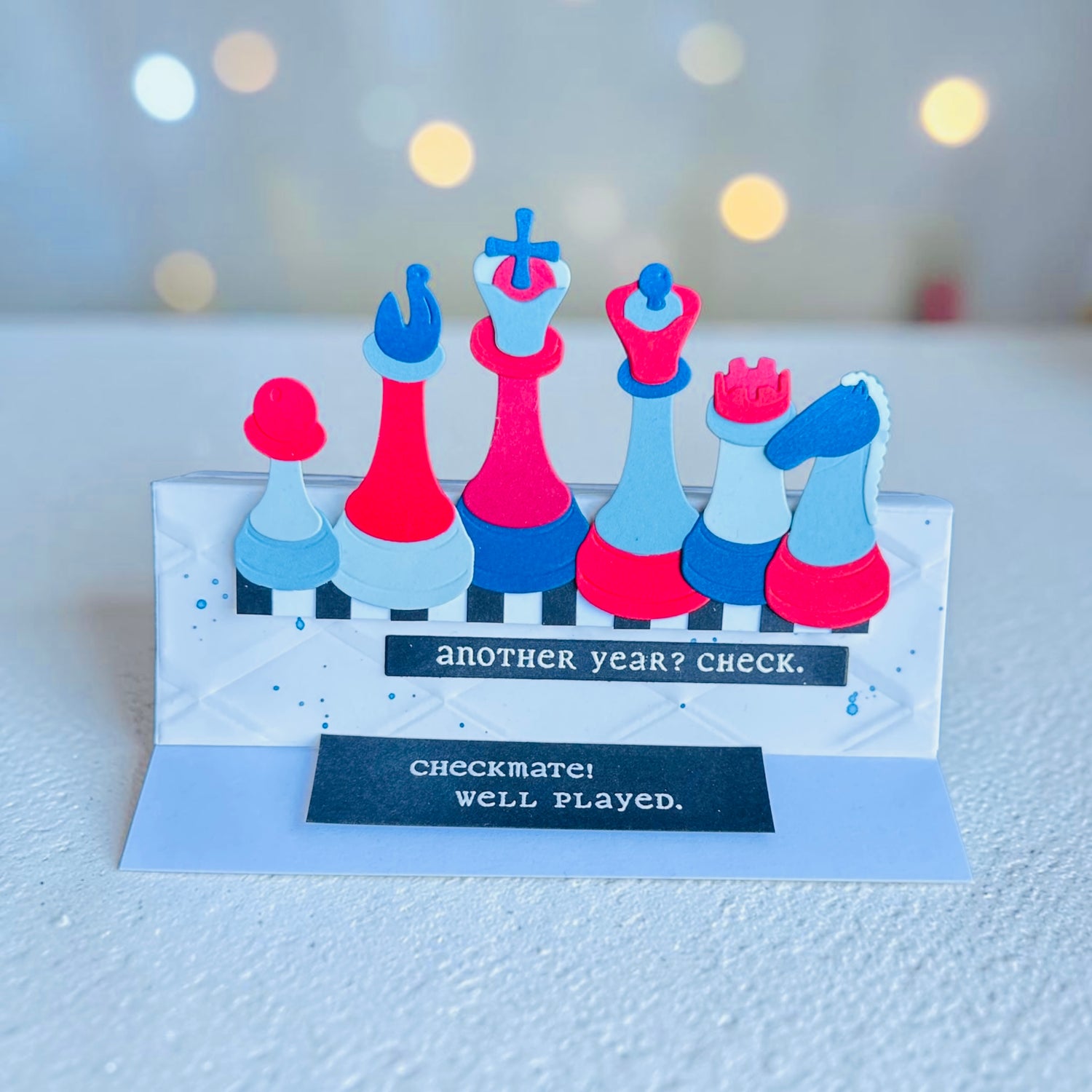 Greeting card with colorful chess pieces and text on a textured surface with blurred lights in the background.