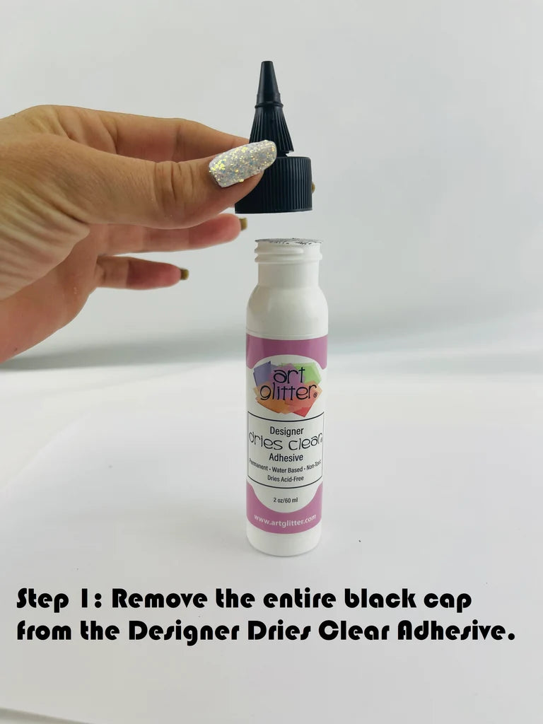 A hand with glittery nail polish holds a small white bottle of Designer Dries Clear Adhesive above its black cap, with a text overlay at the bottom reading 'Step 1: Remove the entire black cap from the Designer Dries Clear Adhesive.'.