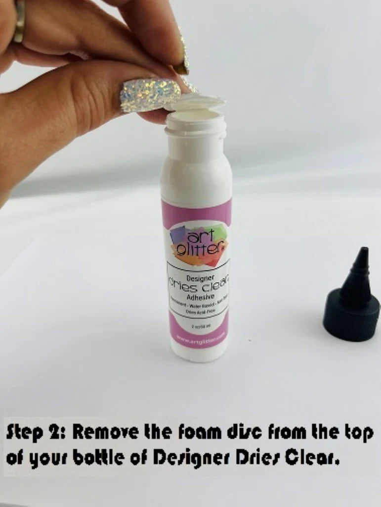 A hand is shown peeling off a clear foam disc from the top of a small white bottle labeled 'art glitter Designer Mac's Clear Adhesive.' The black applicator cap sits to the right on a white surface, with text at the bottom reading 'Step 2: Remove the foam disc from the top of your bottle of Designer Drier Clear.