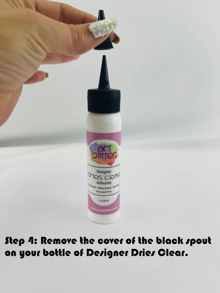A hand with a glittery manicure holds the cap off a small white bottle of Art Glitters Designer dries Clear adhesive. The bottle has a pink label and a black spout cap.