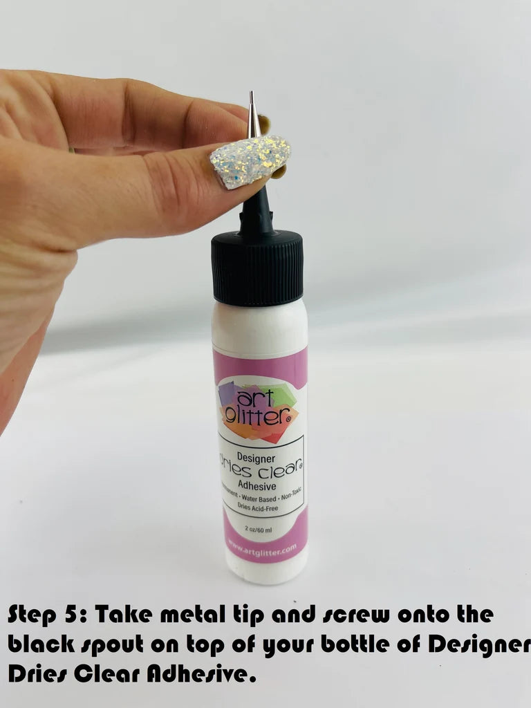A hand with glittery nail polish holds a small bottle of Art Glitter Designer Dries Clear Adhesive. The bottle has a white body, a pink label, and a black cap with a metal applicator tip.