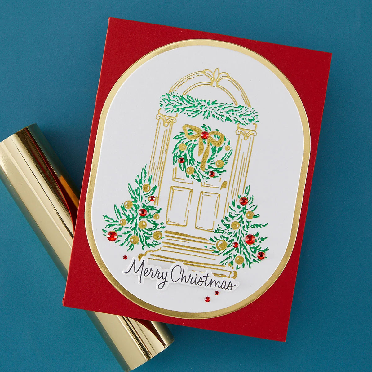 A red Christmas greeting card with a white oval center featuring an illustrated holiday door with green wreaths and gold trim, plus the words 'Merry Christmas' in script. The card is photographed on a blue background with gold cylindrical props partially visible underneath.