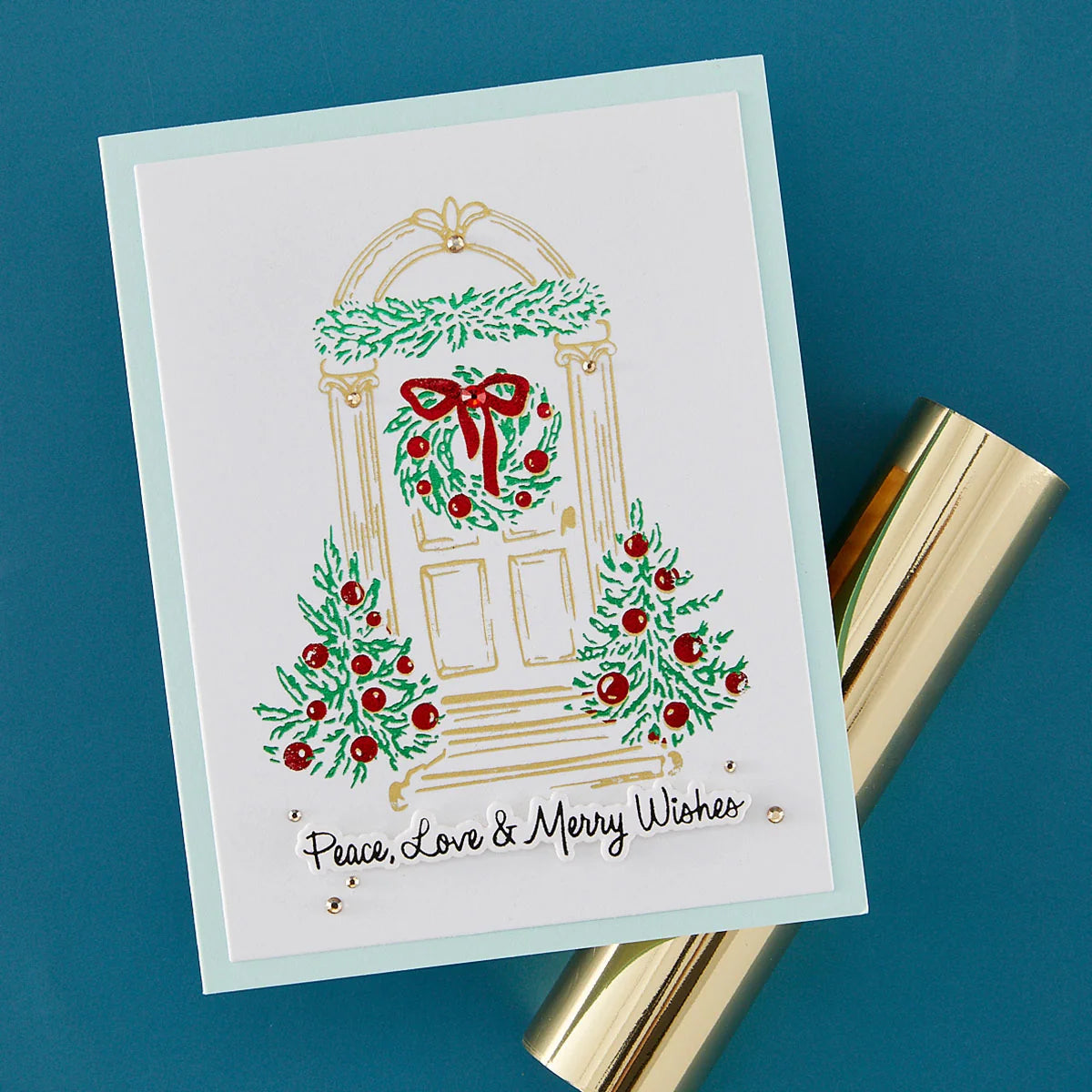 A square white greeting card with a holiday door illustration, green wreaths and garlands, red berries, and the text 'Peace, Love & Merry Wishes,' photographed on a blue background with a gold cylindrical prop beside it.