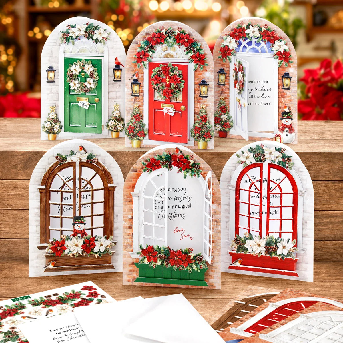 Six arched holiday door and window cards arranged on a wooden surface, each decorated with festive wreaths, lanterns, flowers, and printed holiday phrases. Papers are visible in the foreground, with a softly blurred warm holiday background.