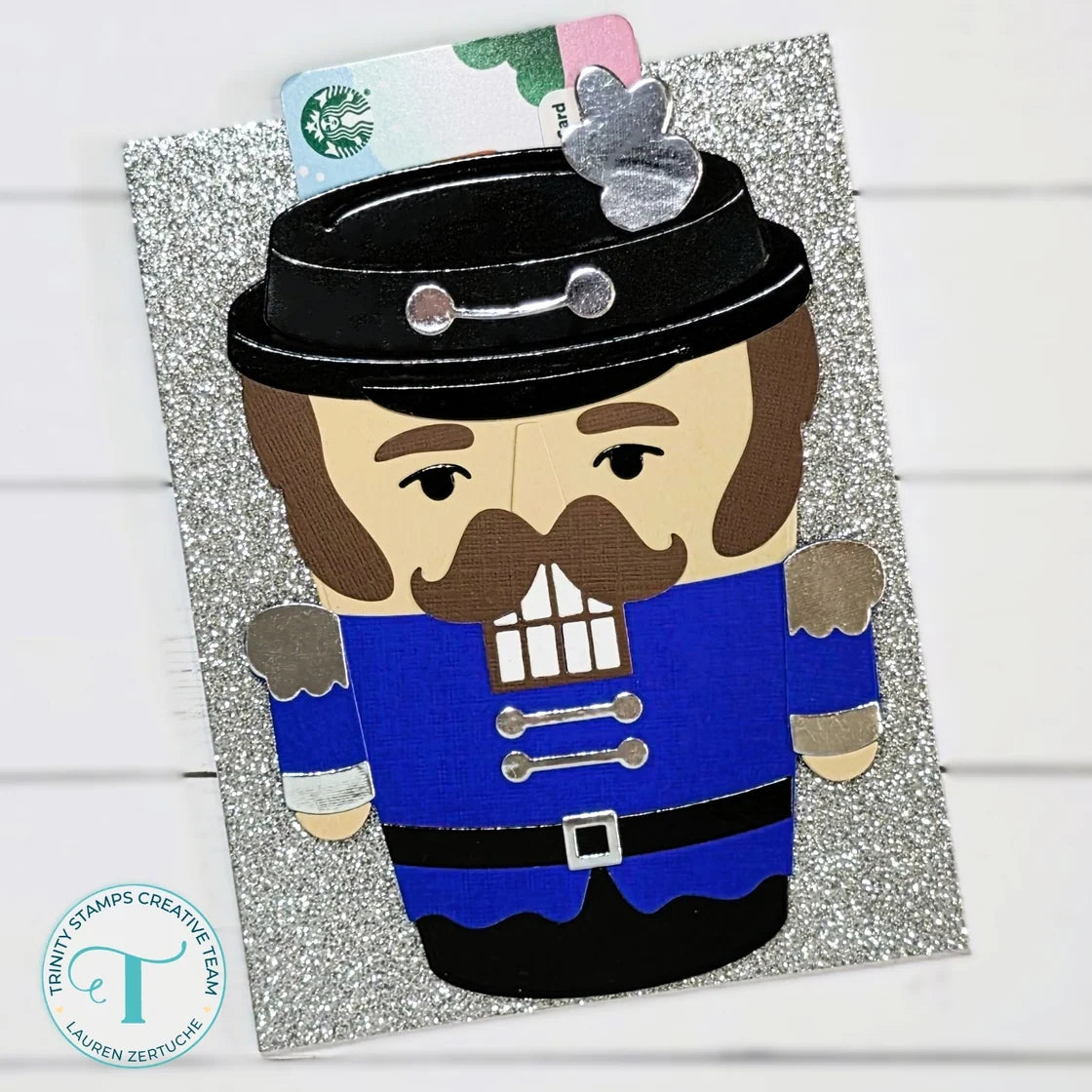A Christmas nutcracker card featuring a paper-cut nutcracker in a blue coat and black hat on a glittery silver background, with a Starbucks card tucked behind the head and a small silver ornament.