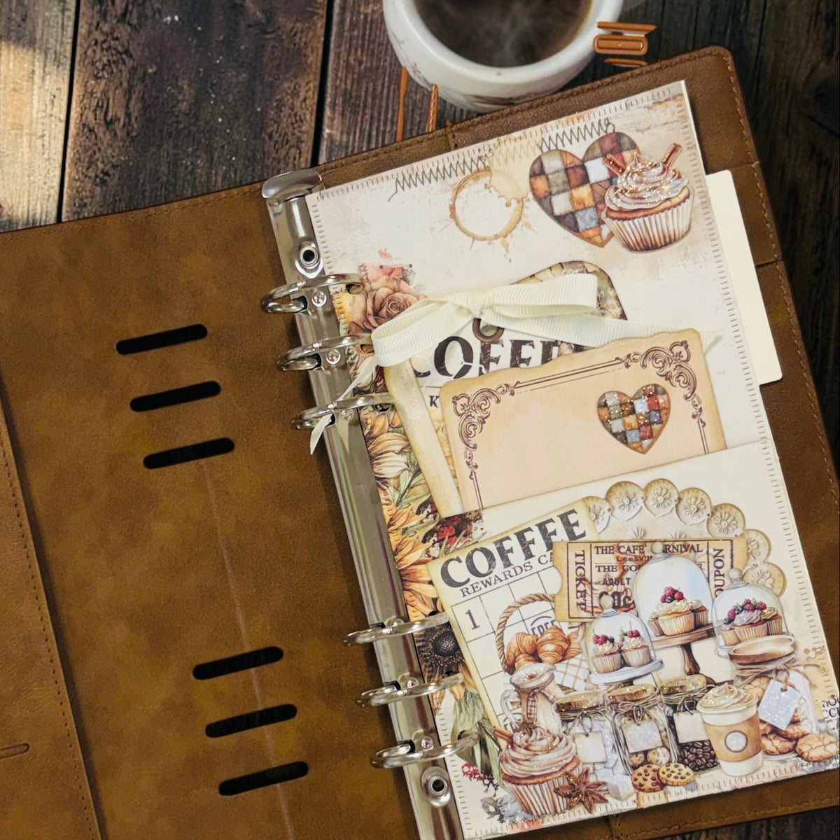 A brown faux-leather ring binder planner or journaling binder shown open on a rustic wooden table, with decorative coffee-themed inserts and a white mug of coffee partially visible at the top right.