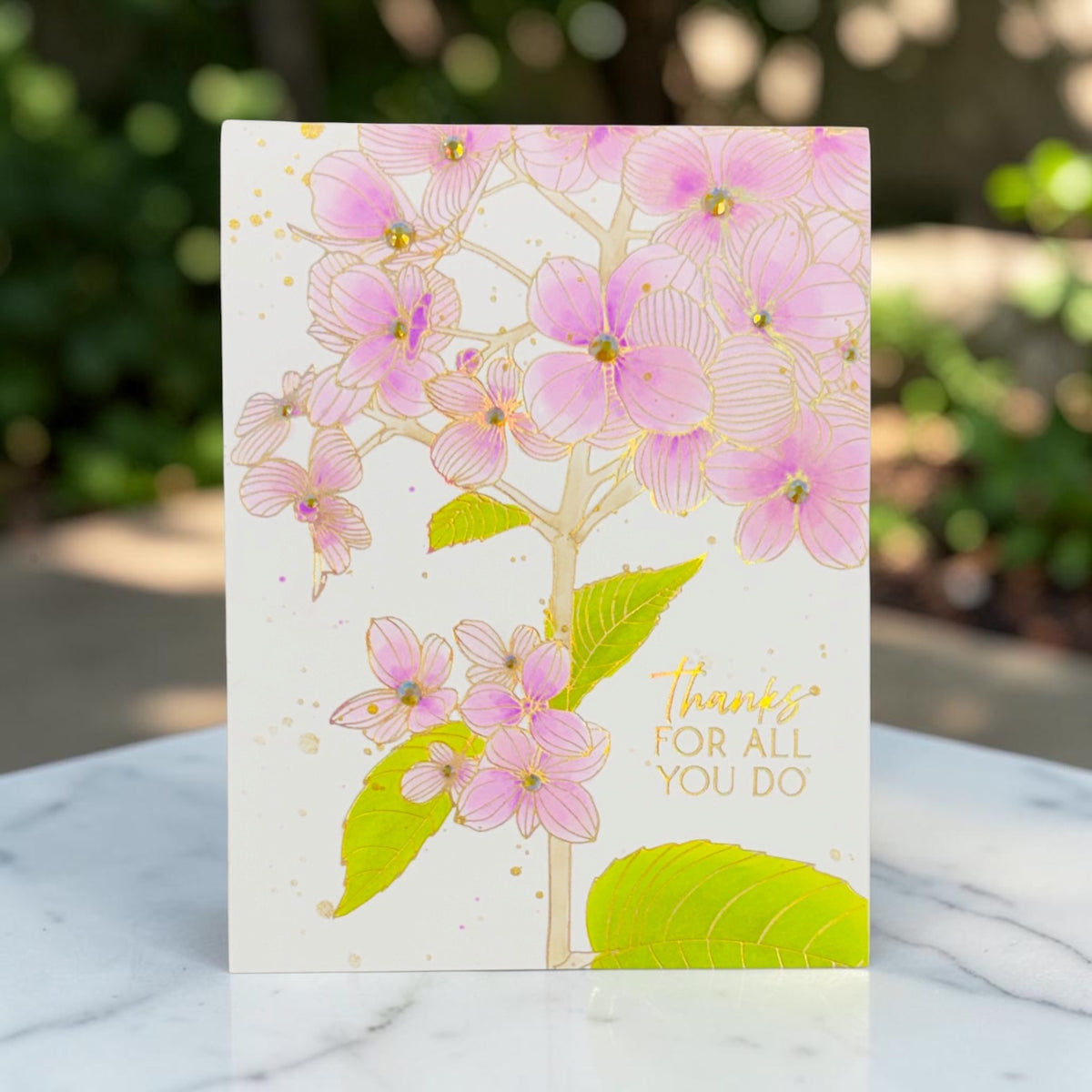 A portrait greeting card featuring a pink hydrangea illustration with gold foil accents and the text 'thanks FOR ALL YOU DO' on the lower right, standing on a white marble surface with a blurred outdoor garden background.