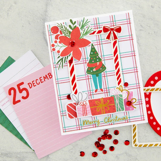 A handmade Christmas greeting card with a festive holiday design, styled on a light gray marble background with stationery props including a pink lined notepad, red rhinestones, and a red polka dot ribbon.
