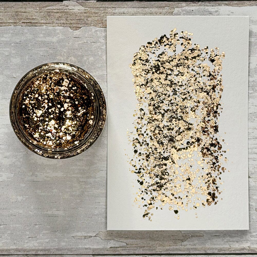 A small glass jar filled with metallic gold and black glitter sits beside a white sheet of paper spread with a matching gold-and-black glitter swatch, photographed on a light gray wood surface.