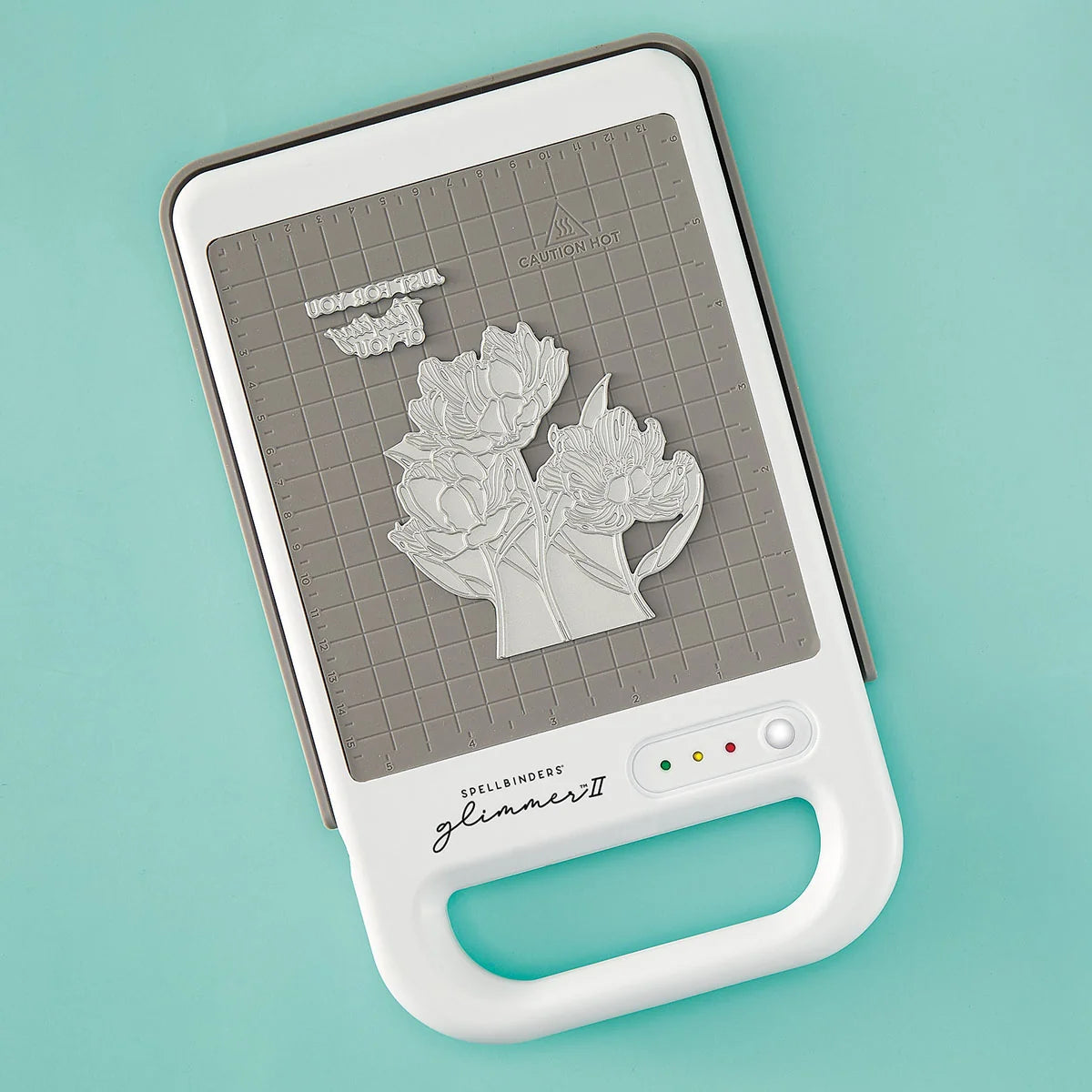 A Spellbinders Glimmer II hot foiling machine out of the box on a light aqua background, featuring a white handle, gray grid cutting mat, and floral press plate.