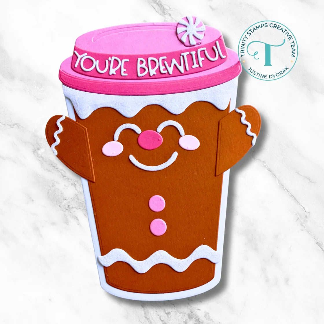 A gingerbread-themed coffee cup craft decoration with a pink lid that reads 'YOU'RE BREENTFUL' and a white circular logo in the upper right on a light marble background.