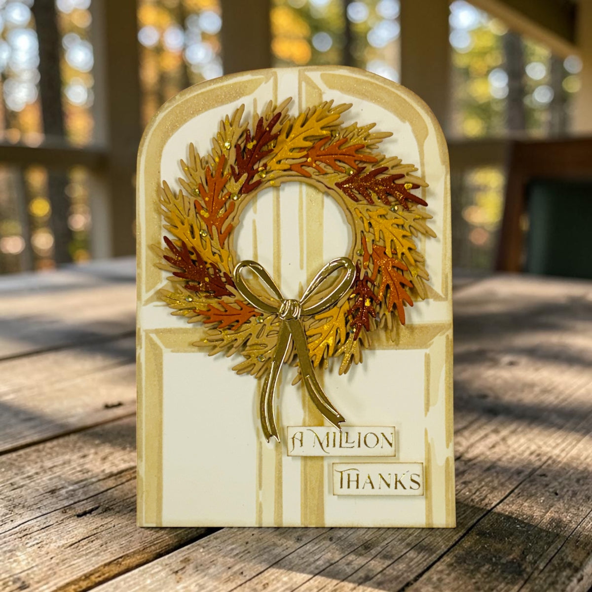A handmade autumn-themed thank you card featuring a 3D leaf wreath with a gold bow on a cream-colored arched background, placed on a rustic wooden table with a blurred outdoor deck background.