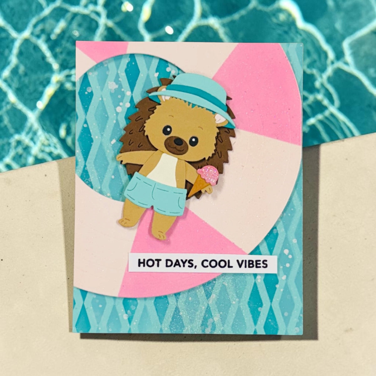 A handmade greeting card featuring a cute cartoon hedgehog wearing a blue hat and shorts, holding an ice cream cone. The card has a pink and white striped background with a turquoise patterned border and the text 'HOT DAYS, COOL VIBES'.