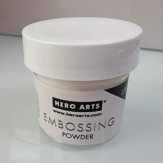 A small white plastic jar of clear Hero Arts Embossing Powder with a white lid, shown on a light reflective surface.