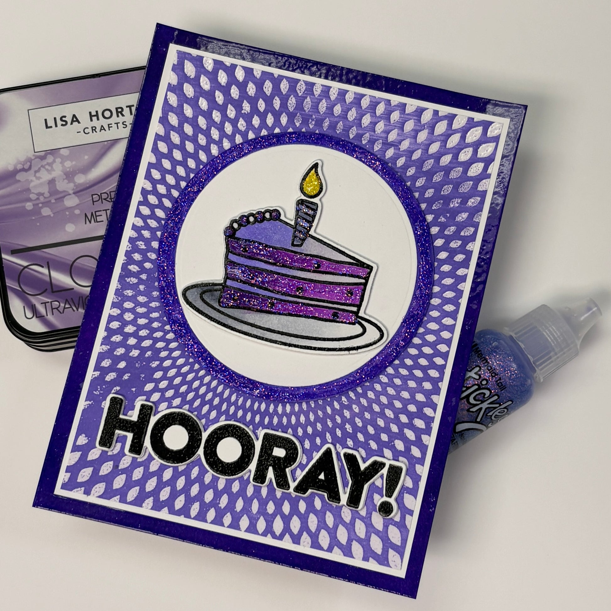 Purple birthday card with cake illustration and 'HOORAY!' text, surrounded by crafting supplies.