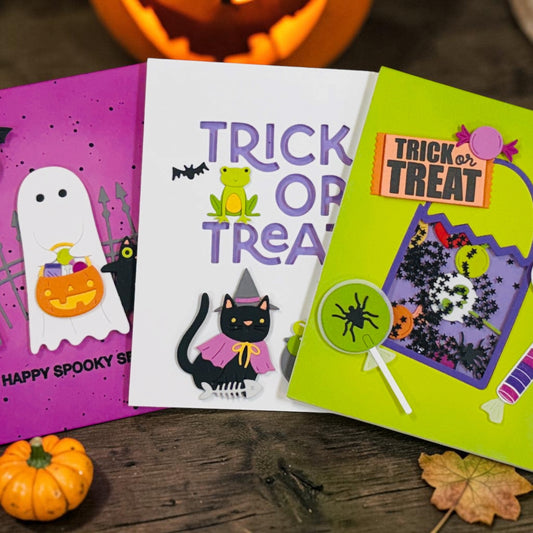 Three colorful Halloween greeting cards arranged on a rustic wooden surface with small pumpkins and a leaf. The cards feature spooky designs including a white ghost on a purple card, a black cat with purple text on a white card, and a haunted house scene on a bright green card.