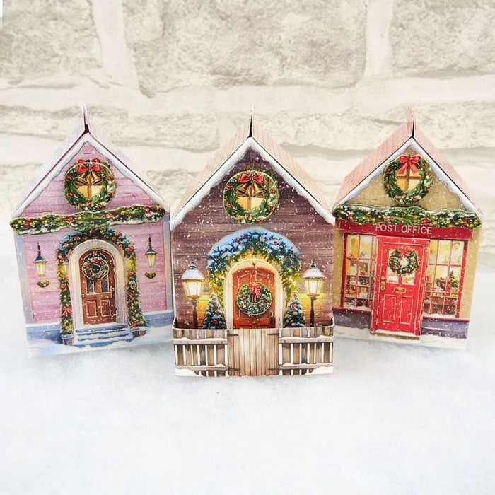 Three small Christmas village house ornaments shaped like a pink house, a brown house, and a red post office, decorated with green garlands, wreaths, and warm glowing lights, displayed against a white brick wall and white surface.