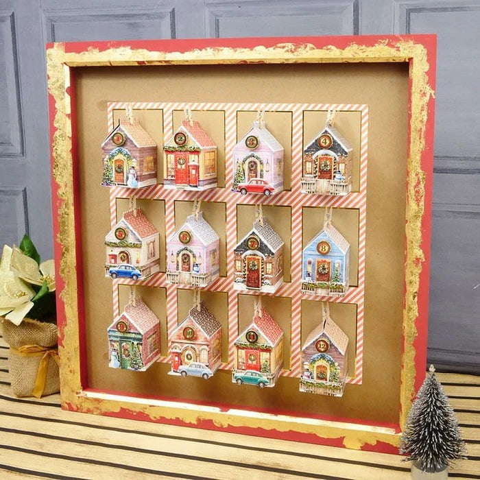 A framed Christmas ornament advent calendar featuring 12 small colorful house ornaments arranged in a 3x4 grid on a kraft paper background, set in a rustic red and gold frame with holiday decorations around it.