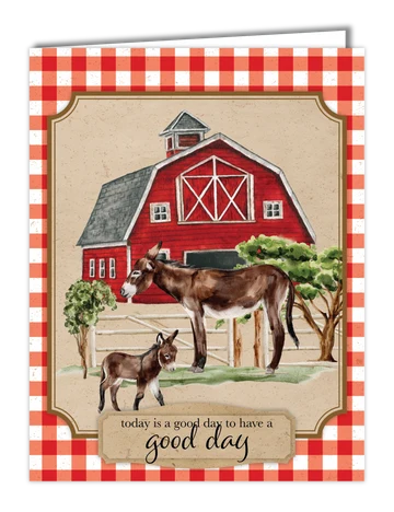 A greeting card featuring a red barn, a standing donkey, and a foal, with the text 'today is a good day to have a good day' on a beige panel. The card has a red and white gingham border and is shown slightly open at the top.