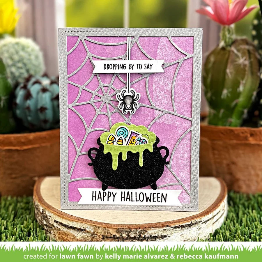 Halloween-themed card with a cauldron and spider on a pink background, surrounded by plants.