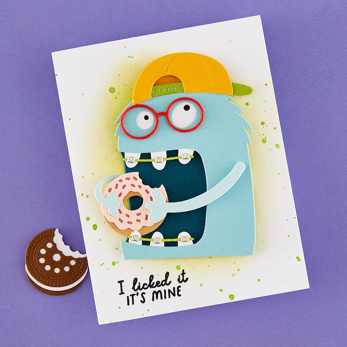 A handmade greeting card featuring a blue monster character wearing red glasses and a yellow cap, holding a pink frosted donut. The card has green paint splatters, the text 'I licked it IT'S MINE' at the bottom, and is displayed on a purple background next to a cookie cutout.