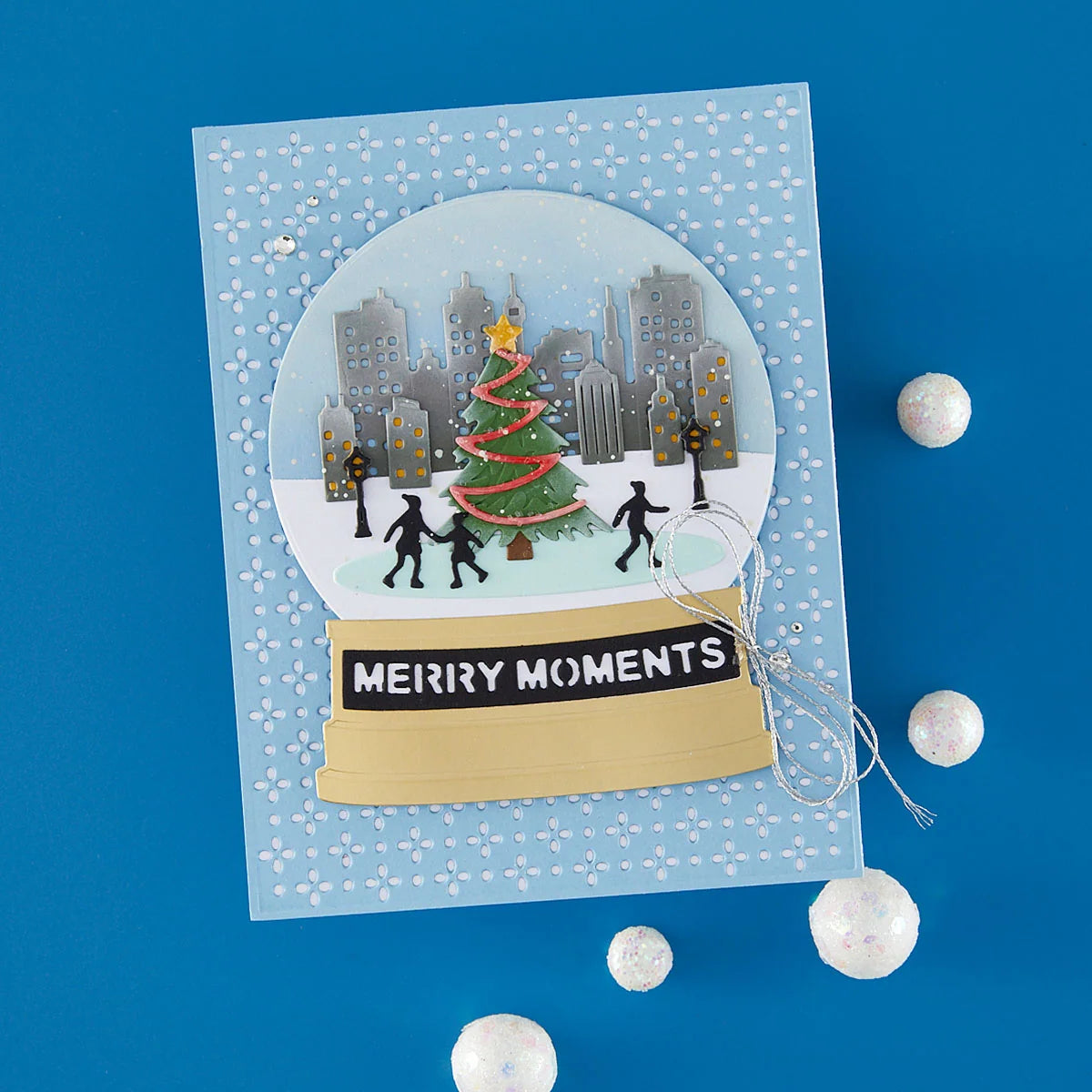 A handmade layered card featuring a circular snow globe design with a Christmas tree, city skyline, and silhouettes of people, set on a light blue patterned background with a gold banner reading 'MERRY MOMENTS'. The card is displayed on a solid blue background with decorative white pom-poms.