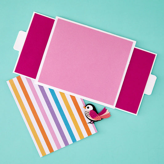 A pink rectangular card or envelope-style paper item with magenta side flaps, laid on a turquoise background, with a striped paper sheet and a small bird embellishment peeking out from underneath.