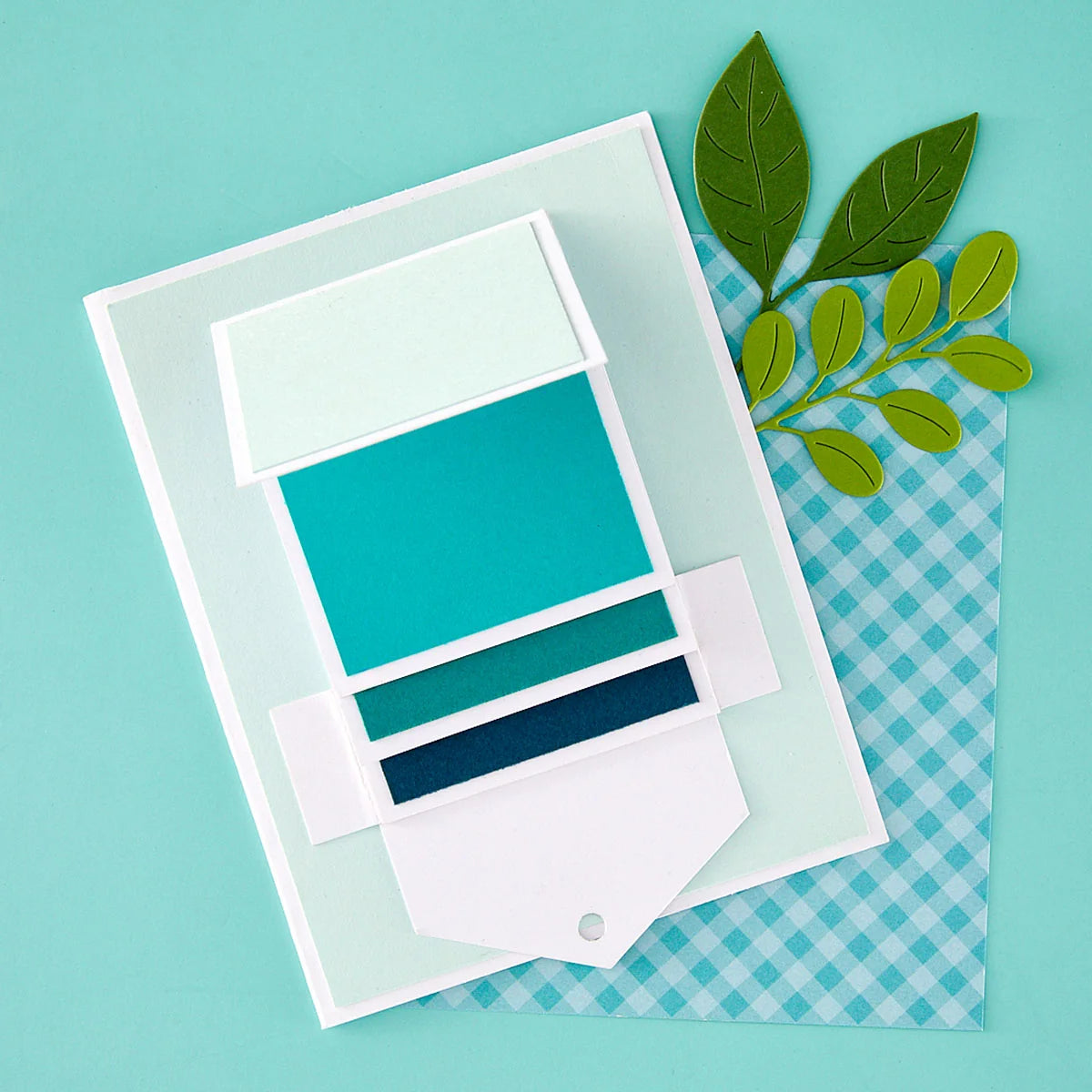 A layered paper craft or card design with white cardstock shapes and teal, green, and navy colored sections, placed on a turquoise background with a blue-and-white gingham sheet and green leaf accents.