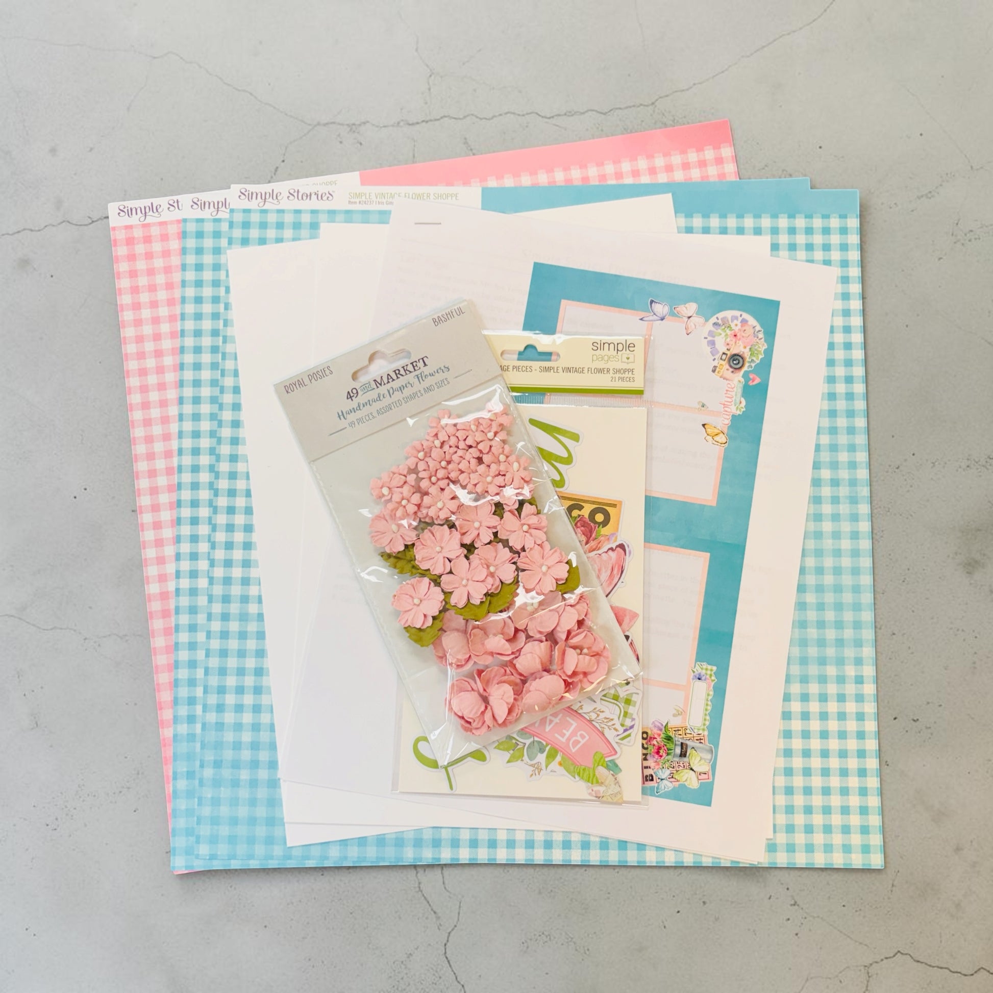 A flat-lay of craft supplies on a light gray surface, featuring a package of pink floral embellishments layered over patterned paper sheets in pink gingham, light blue gingham, and white.