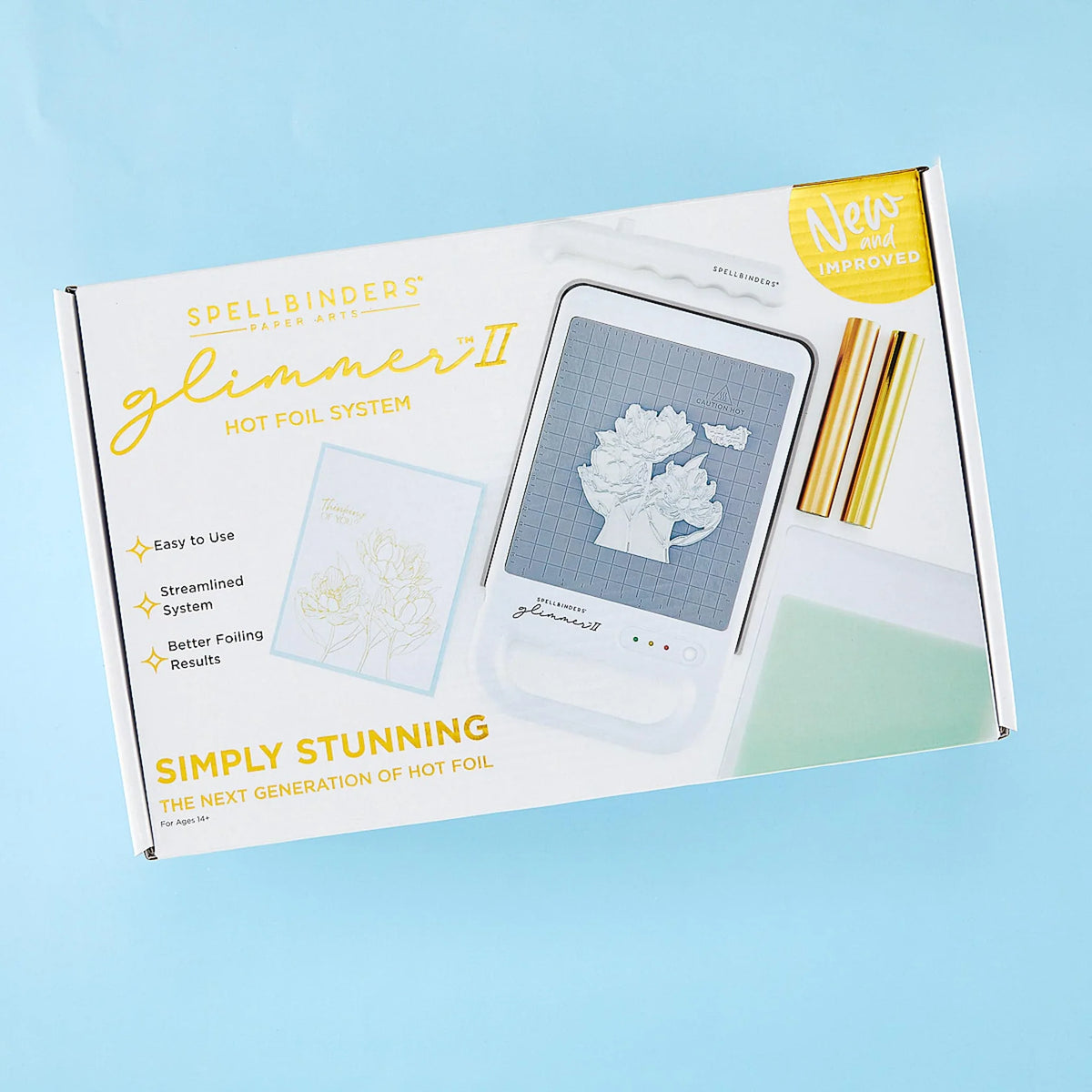 Spellbinders Glimmer II Hot Foil System kit in its retail box, displayed on a light blue background. The white box features gold lettering and showcases the included hot foil machine, and gold metallic rolls of foil.