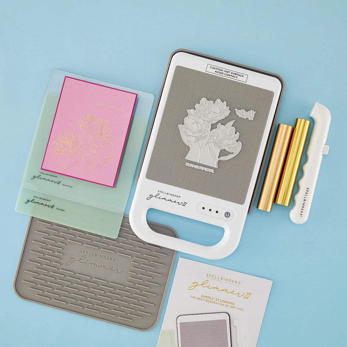 Spellbinders Glimmer II foiling supplies arranged on a light blue background, including a white and gray machine, gold rolls of foil, a white magnetic tool, a gray silicone mat, a pink foiled card panel, and an instruction booklet.