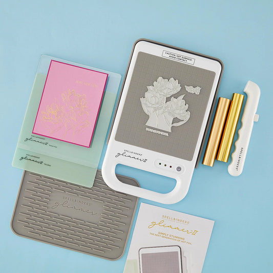 Spellbinders Glimmer II foiling supplies arranged on a light blue background, including a white and gray machine, gold rolls of foil, a white magnetic tool, a gray silicone mat, a pink foiled card panel, and an instruction booklet.