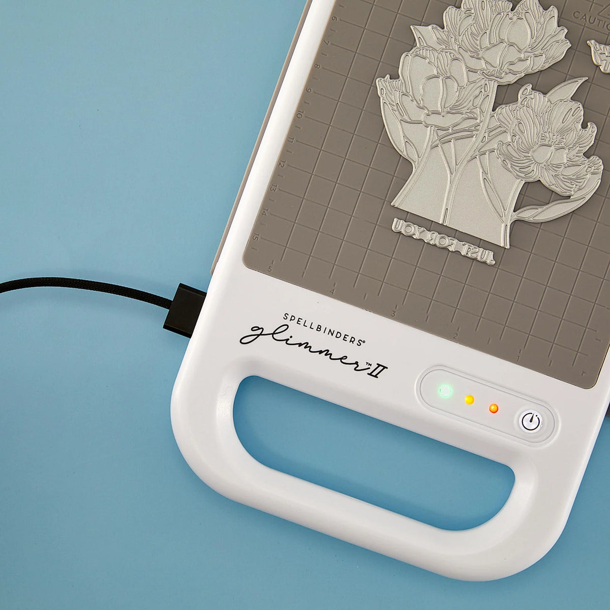 A close-up of a Spellbinders Glimmer II hot foiling machine on a light blue background. The white body features a handle, a gray textured  grid heating plate with a floral press plate, and a power button with LED indicators.