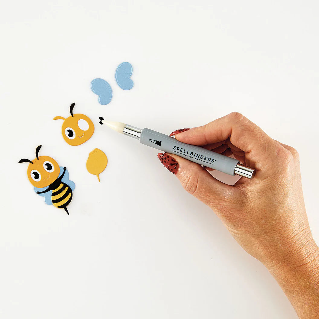 Hand holding a spellbinders tool with cartoon bee and butterfly illustrations on a white background