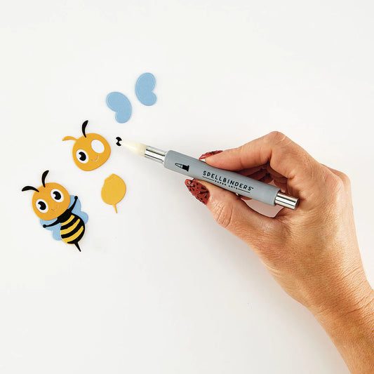 Hand holding a spellbinders tool with cartoon bee and butterfly illustrations on a white background