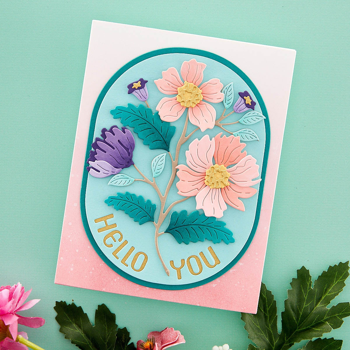 A handmade layered card featuring a 3D floral design with pink and purple flowers, teal leaves, and gold foil text reading 'HELLO YOU' on a light blue oval, set against a pink backing card on a mint green background.